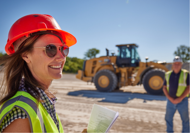 A male and a female Foley employee wearing protective glasses and hard hats stand before a Caterpillar vehicle.
