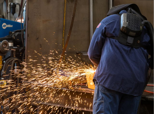 A man wearing a safety mask grinds a piece of metal, making sparks fly.