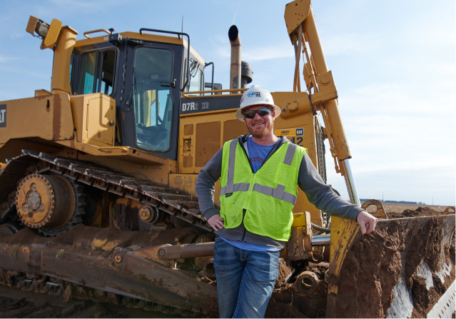 A male Foley wearing a hard hat stands before a large Caterpillar vehicle.