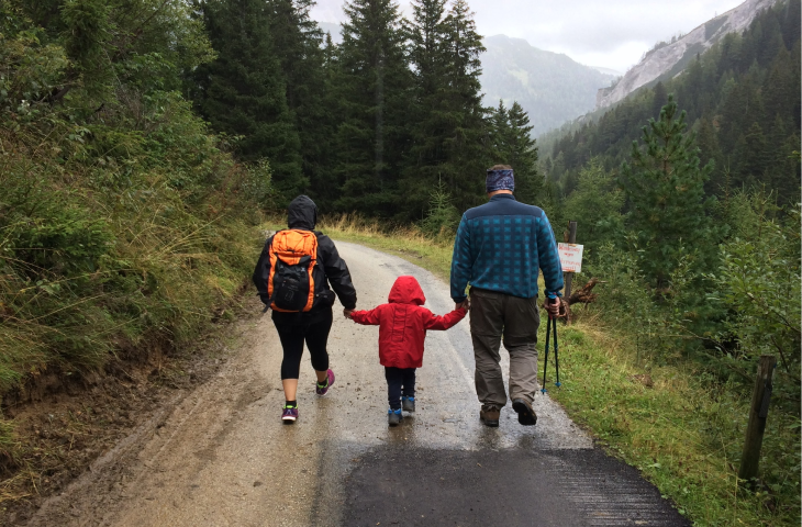 A mother and father hold their child's hands as they walk together down a forest track